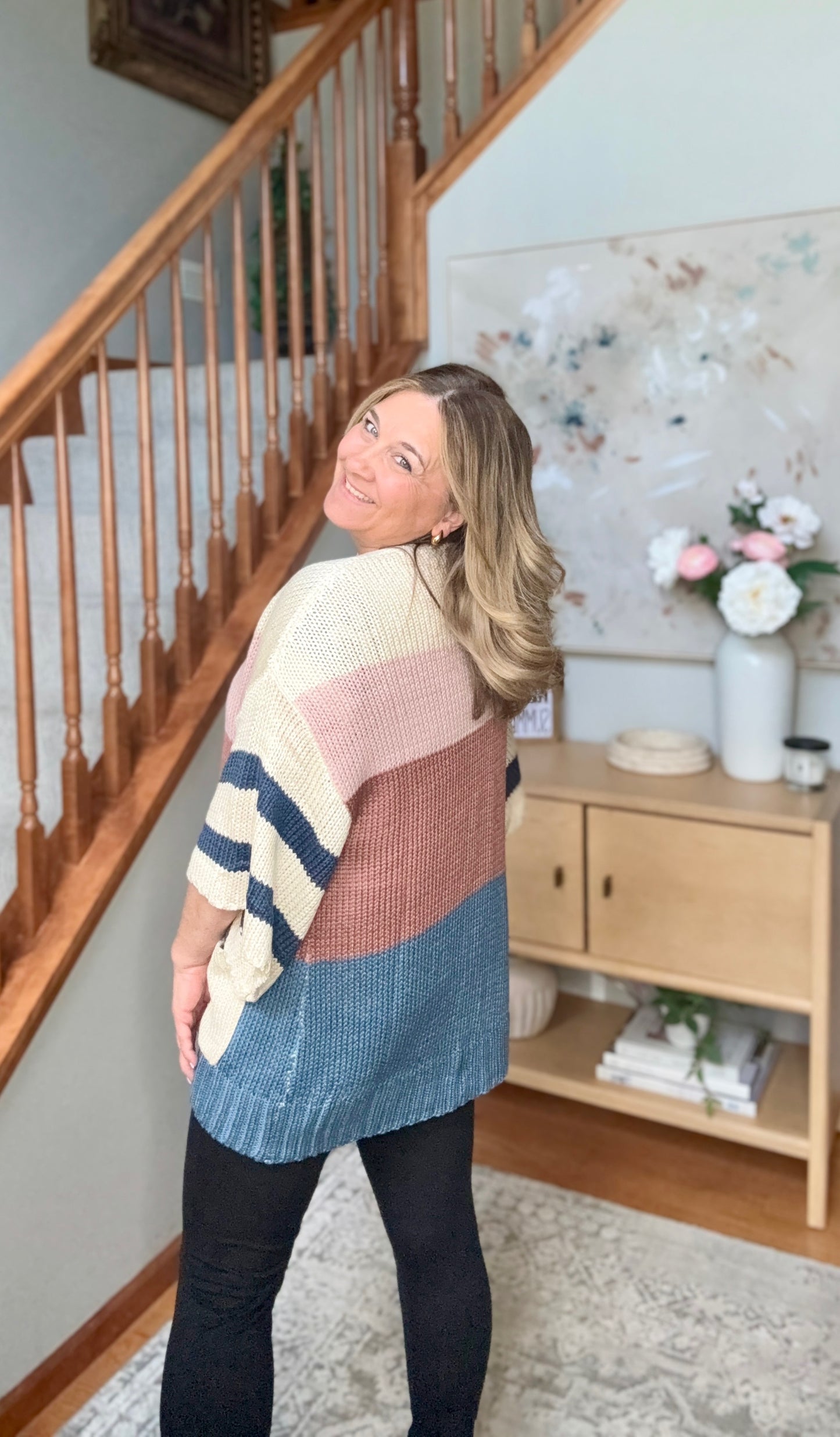 A woman wearing a color-block knit sweater with cream, dusty pink, and blue sections, standing in a home entryway near a wooden staircase.