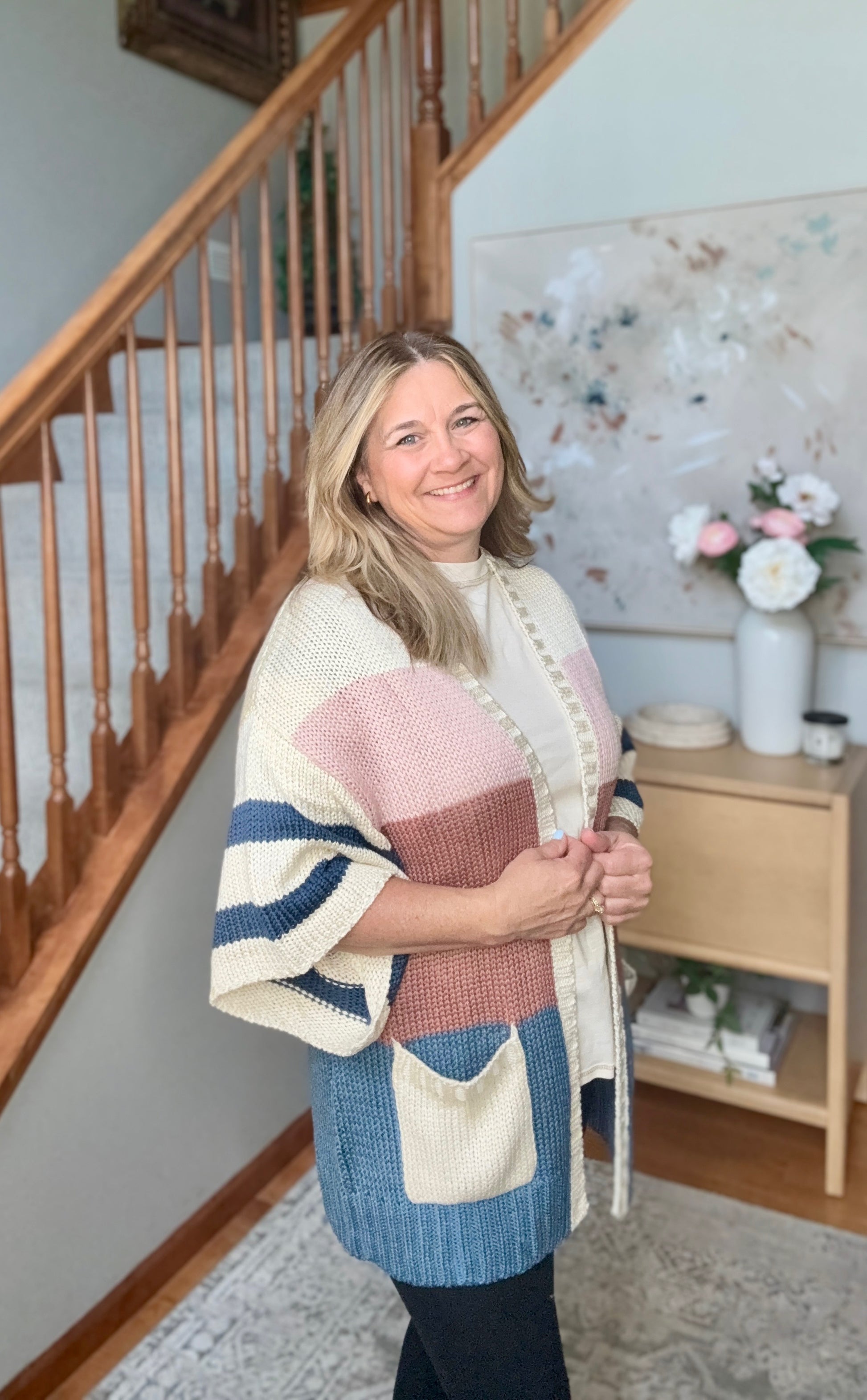 A woman wearing a chunky knit color-block poncho sweater with cream, pink, blue, and navy panels, standing in a home interior next to a wooden staircase.