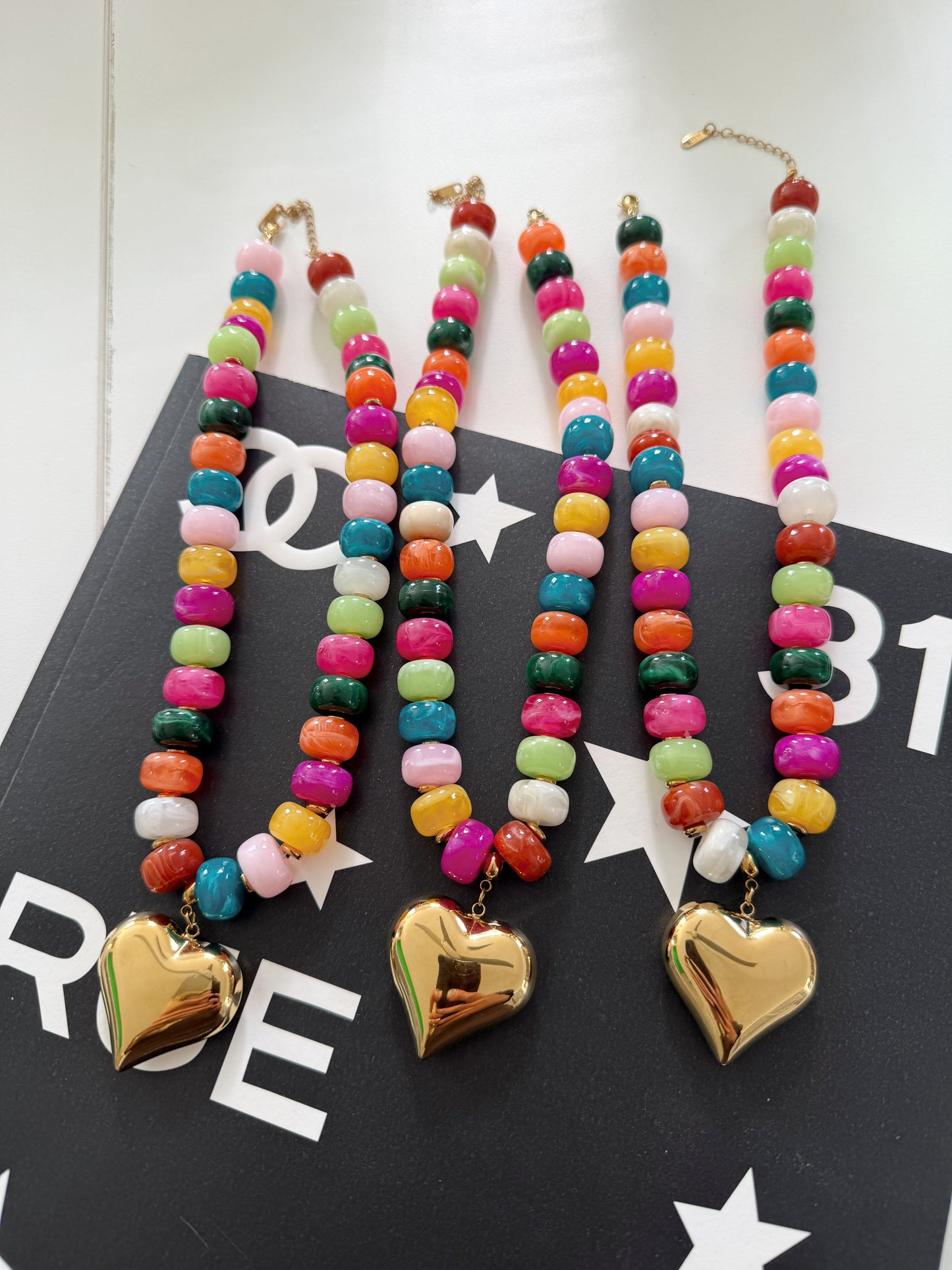 Three colorful beaded necklaces with gold heart pendants, displayed on a black surface with white star and logo patterns.