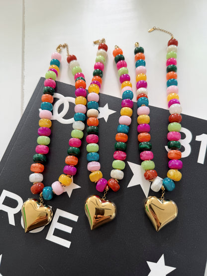 Three colorful beaded necklaces with gold heart pendants, displayed on a black surface with white star and logo patterns.