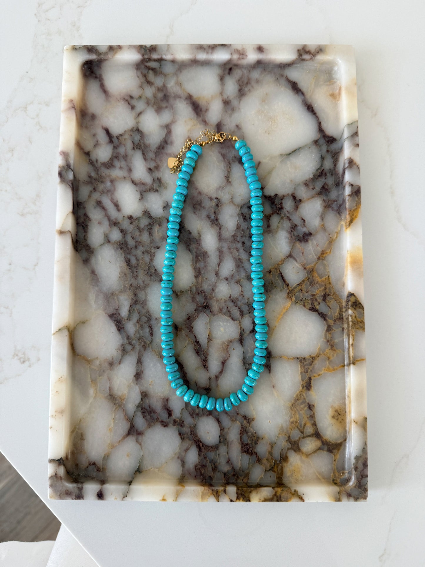 A turquoise beaded necklace with small round beads and a gold clasp, displayed on a rectangular white and gray marbled tray.
