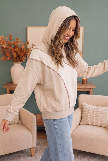A woman wearing a beige zip-up hoodie with diagonal contrast stitching and light blue jeans, standing in a cozy living room setting with beige armchairs and a green wall.
