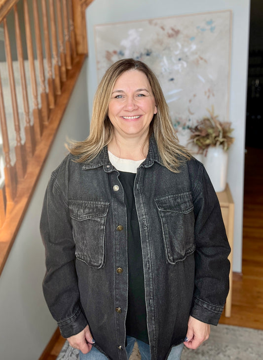 Woman wearing a dark denim jacket in a home setting with a staircase and decorative items in the background.