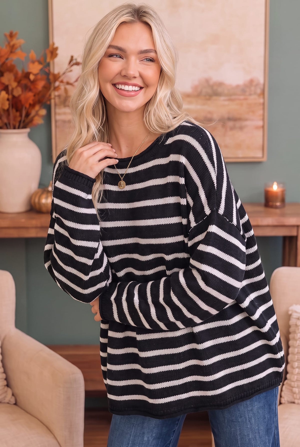 A smiling blonde woman wearing a black and cream horizontal striped knit sweater with blue jeans, posed in a cozy living room setting.