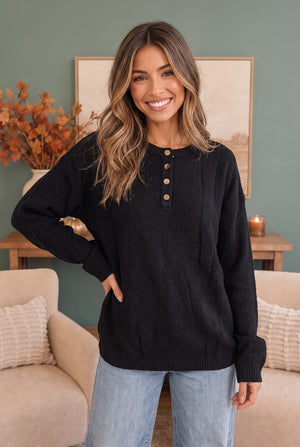 A woman wearing a black textured knit sweater with a three-button henley neckline, paired with light blue jeans, standing in a cozy living room setting.
