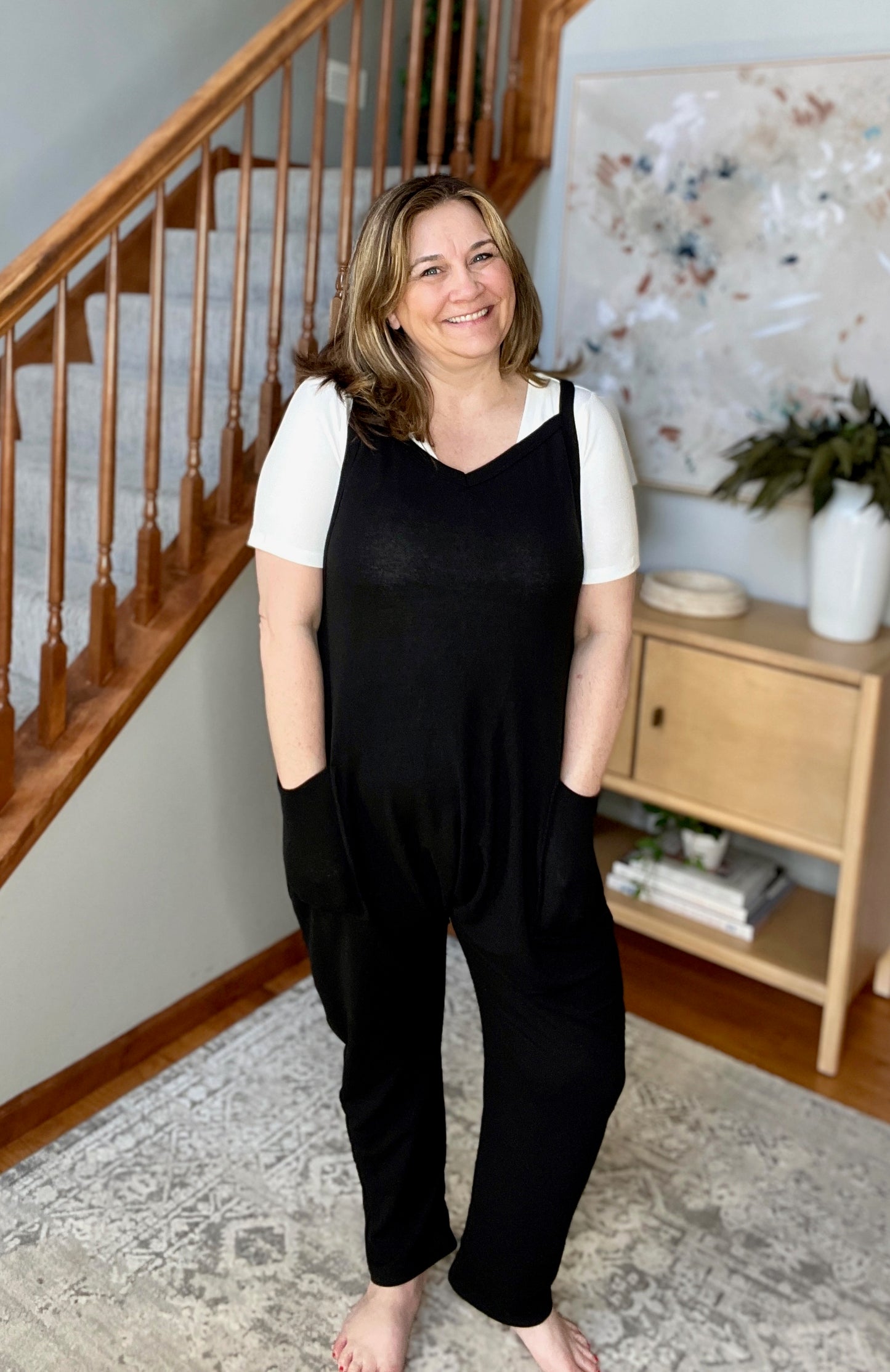 Woman wearing a black jumpsuit standing in a home setting with a staircase and decor.