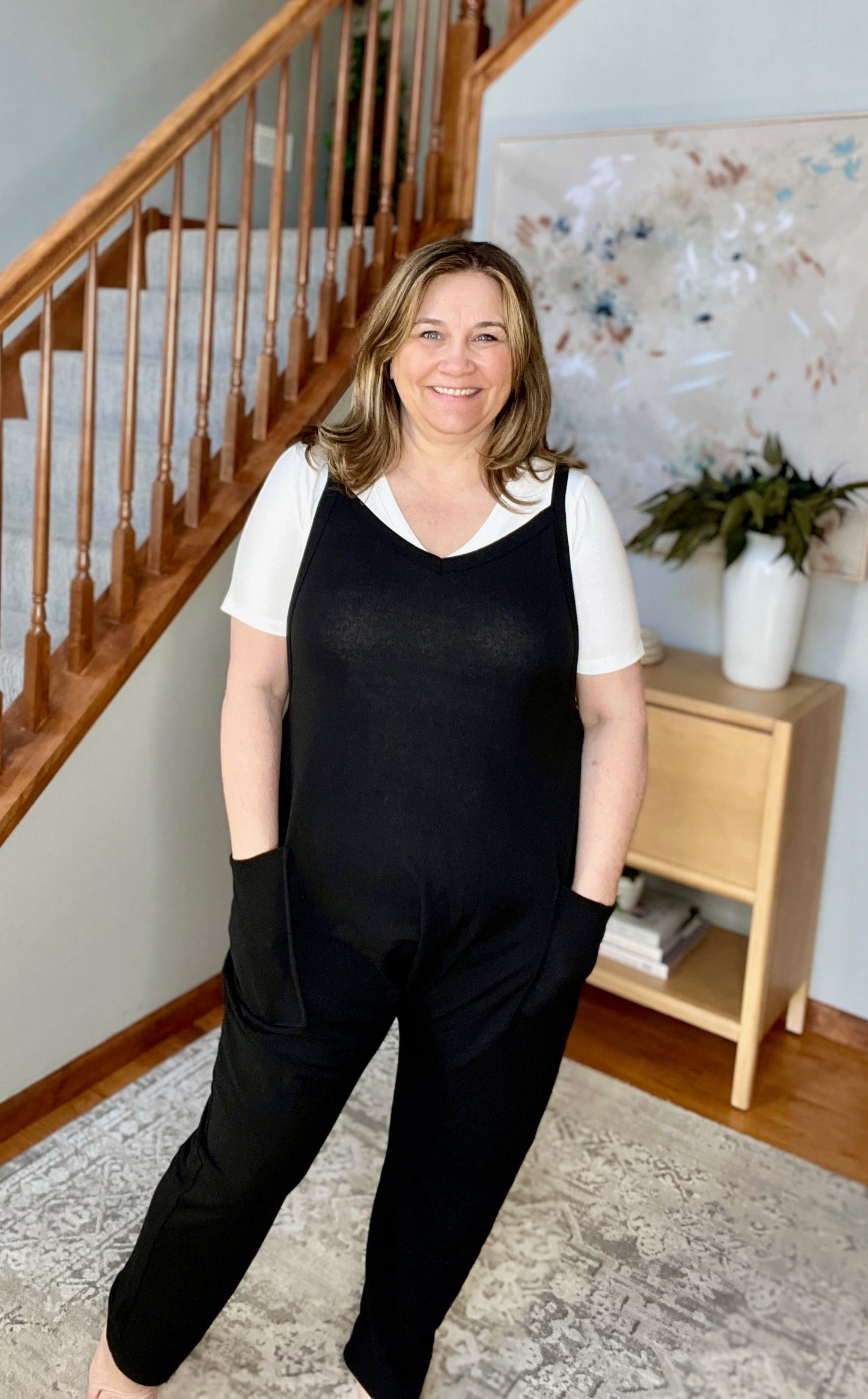 Woman wearing a black jumpsuit standing in a home setting with a staircase and plant in the background.