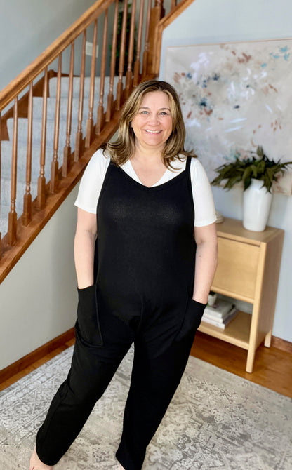 Woman wearing a black jumpsuit standing in a home setting with a staircase and plant in the background.