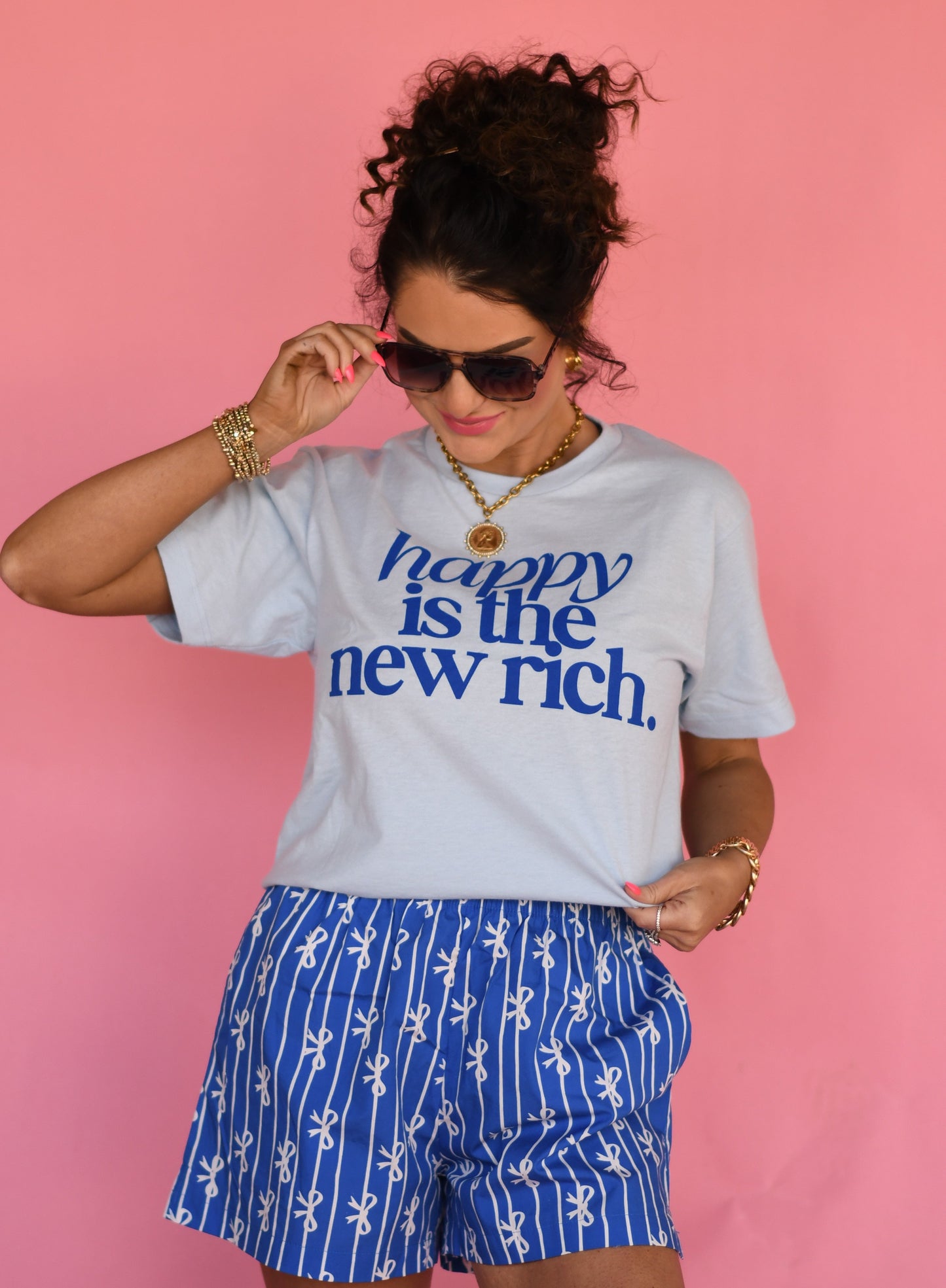 Woman wearing a white t-shirt with 'happy is the new rich' text and blue shorts on a pink background