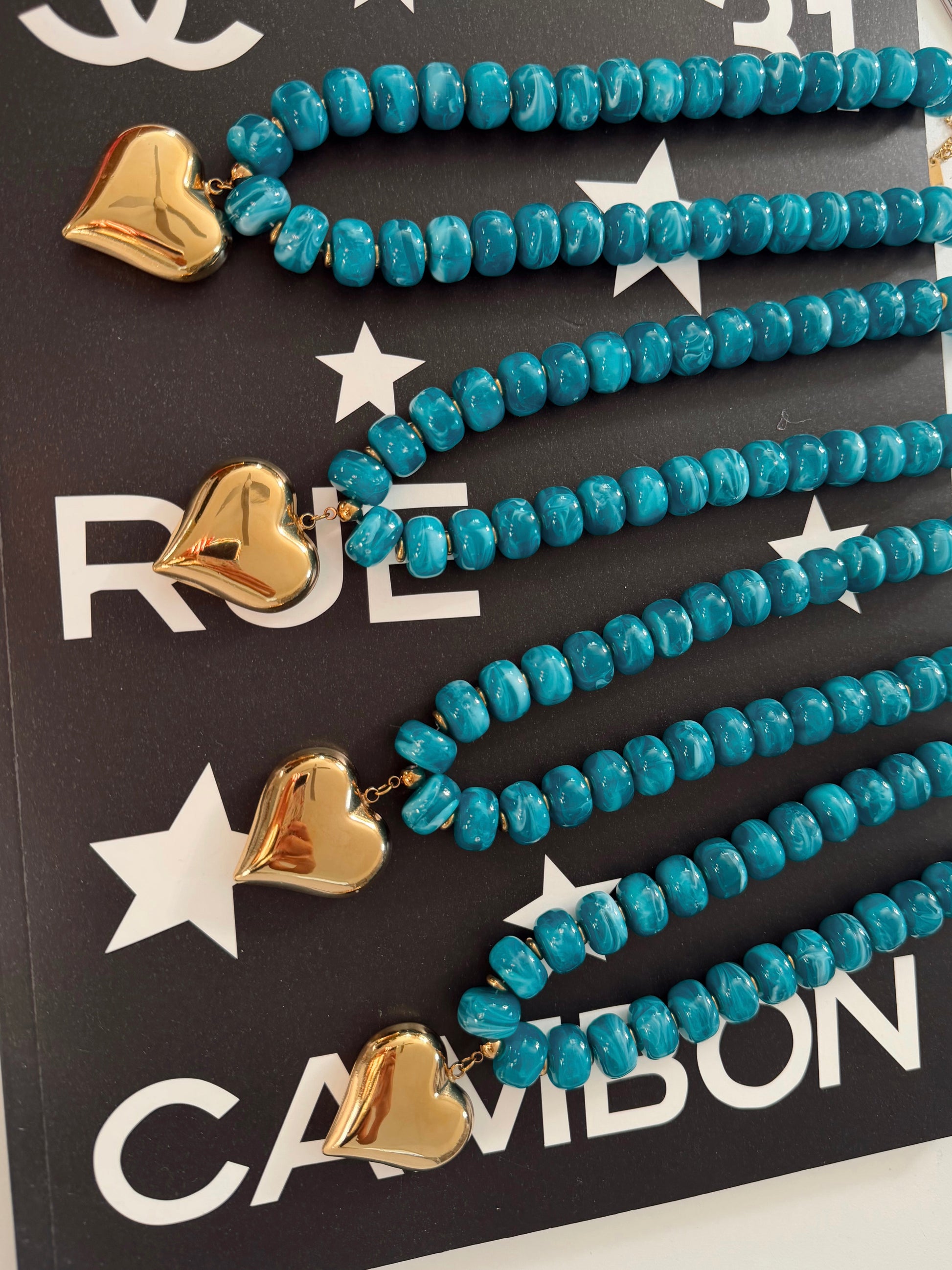 Three turquoise beaded necklaces with gold heart charms displayed on a black card with white stars and the brand name CAMIBON.
