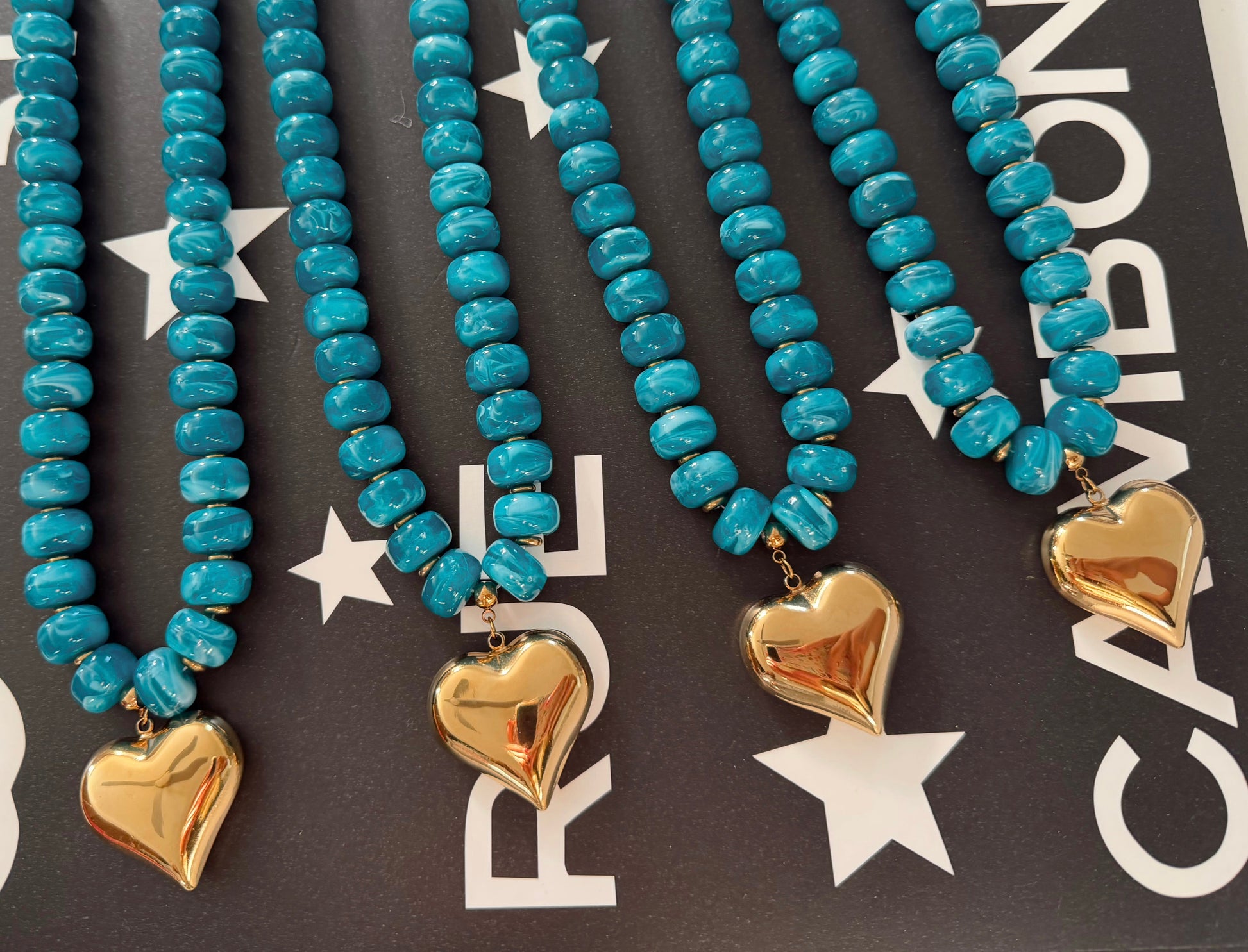 Four turquoise beaded necklaces with gold heart charms, arranged diagonally on a dark background with white star and letter graphics.