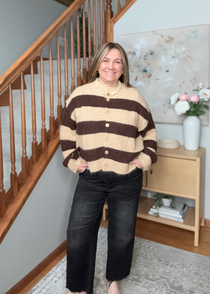 A woman wearing a fuzzy knit cardigan with horizontal brown and beige stripes, paired with black wide-leg jeans, standing in a home interior next to a wooden staircase.