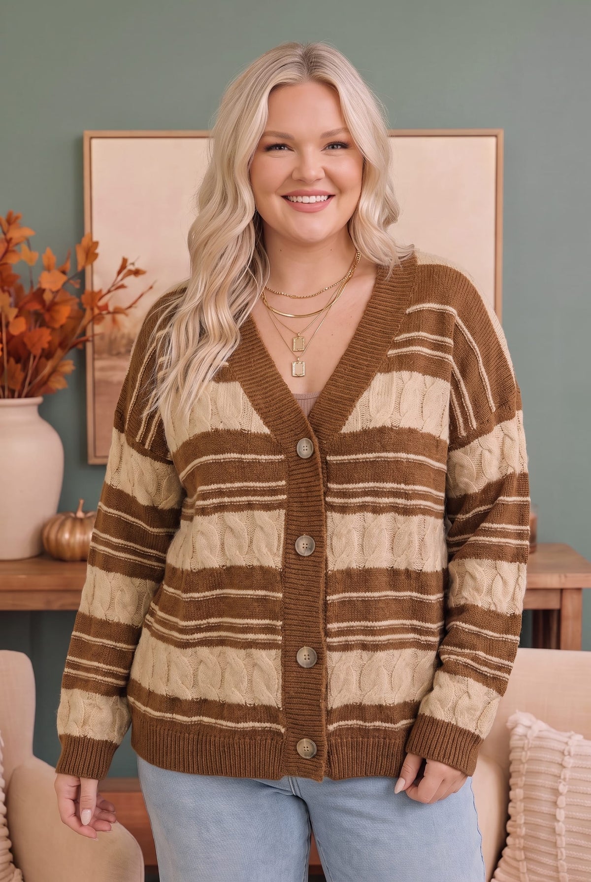 A smiling blonde woman wearing a brown and cream striped cable-knit cardigan sweater with light blue jeans, standing in a cozy room setting.
