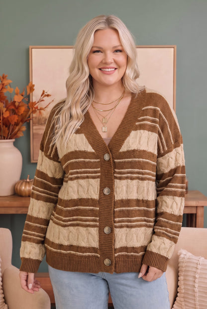 A smiling blonde woman wearing a brown and cream striped cable-knit cardigan sweater with light blue jeans, standing in a cozy room setting.