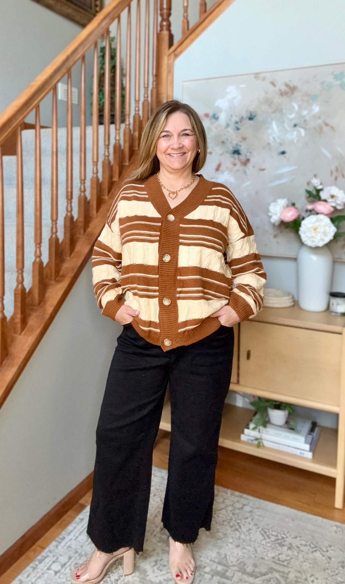 A woman stands in a home interior wearing a brown and cream striped knit cardigan with buttons, paired with black wide-leg pants and beige heeled sandals. A staircase, wood cabinet, and floral decor are visible in the background.