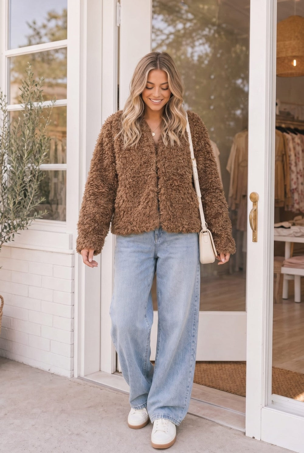 A woman stands in front of a white storefront doorway wearing a brown fuzzy cropped jacket, light blue wide-leg jeans, white sneakers, and carrying a small cream shoulder bag.