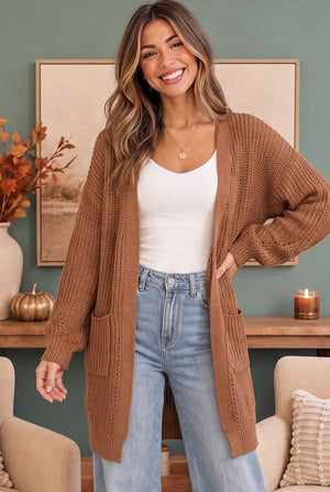 A woman wearing a long, open-front chunky knit cardigan in a warm camel brown, styled over a white tank top and light blue jeans, standing in a cozy room setting.