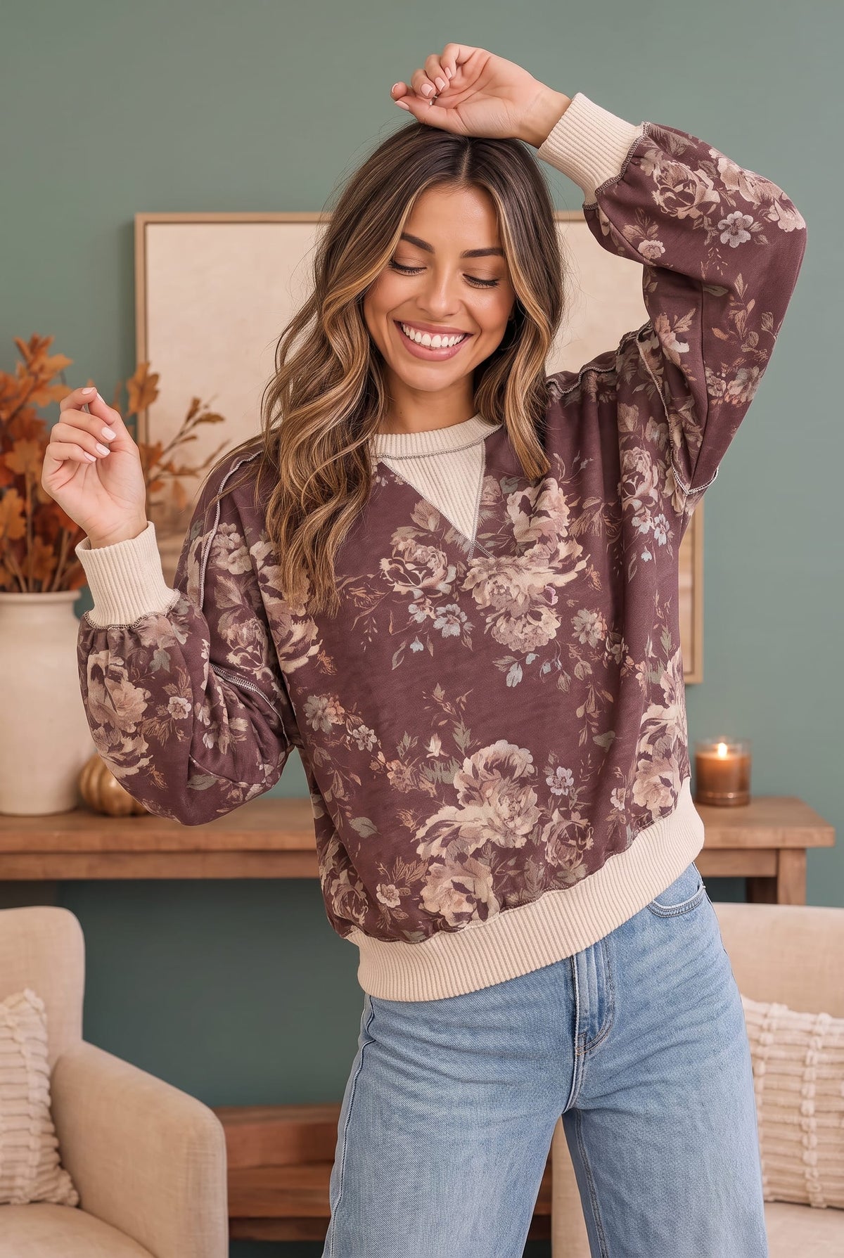 A woman wearing a brown floral long-sleeve sweatshirt with cream ribbed trim, paired with light blue jeans, standing in a cozy living room setting.
