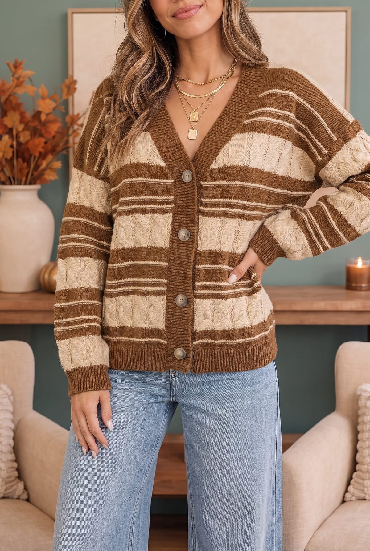 A woman wearing a brown and cream striped cable-knit cardigan sweater with a button-front closure, paired with light blue jeans, in a cozy living room setting.