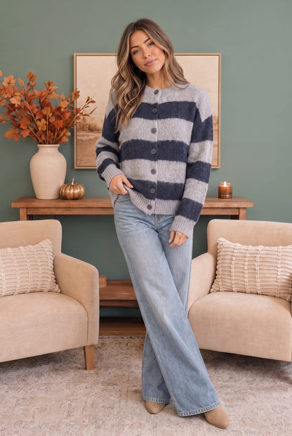 A woman stands in a styled living room wearing a fuzzy knit cardigan with wide navy and light gray horizontal stripes, paired with light-wash flare jeans and tan flats.