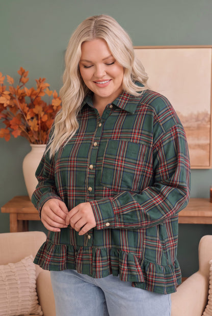 A woman wearing a green plaid button-down shirt with red and white stripes, featuring a ruffled hem and chest pocket, paired with light blue jeans in a cozy indoor setting.