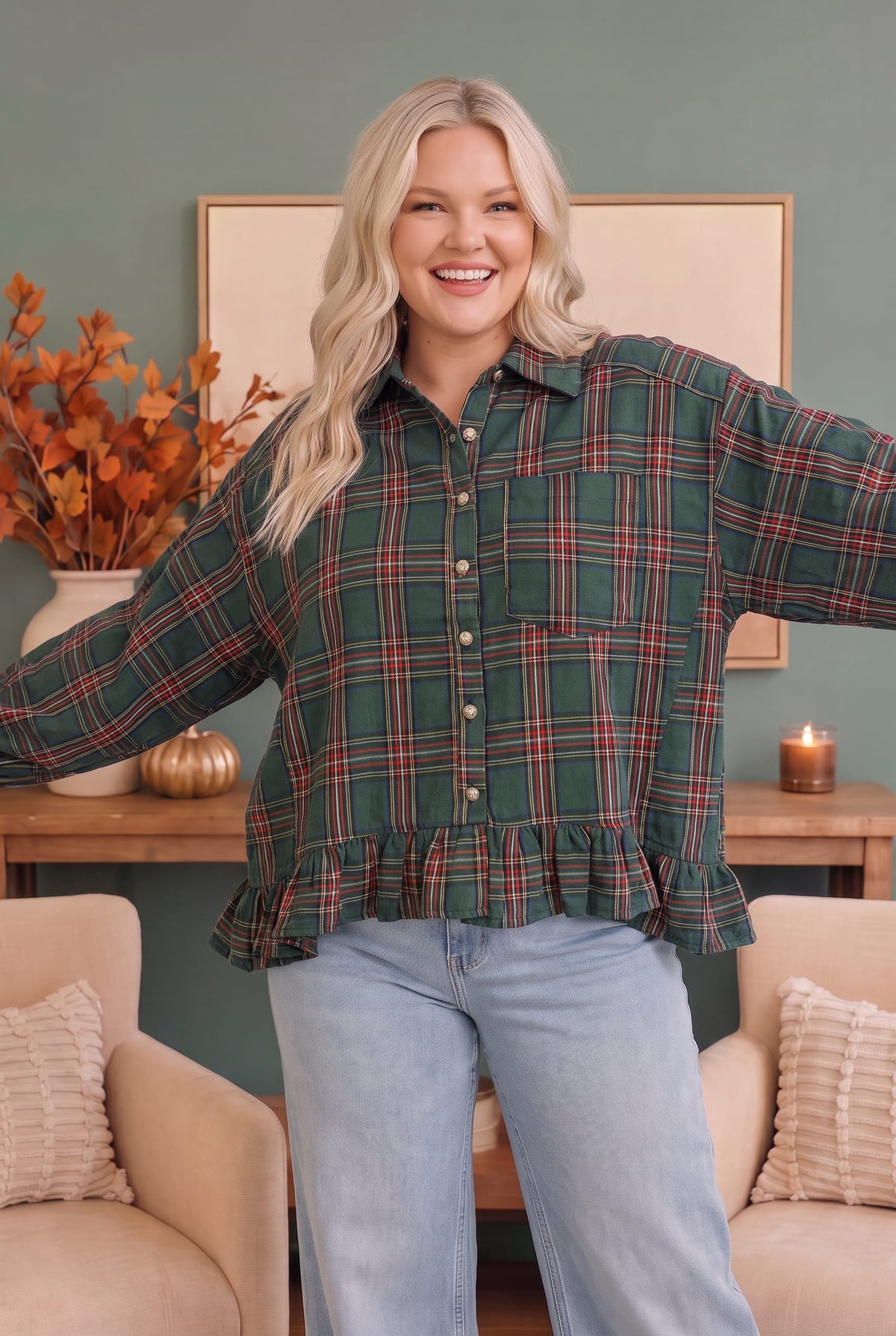 A woman modeling a green plaid button-up shirt with a ruffled hem and light blue jeans, standing in a cozy living room setting with beige armchairs and a wooden console table.