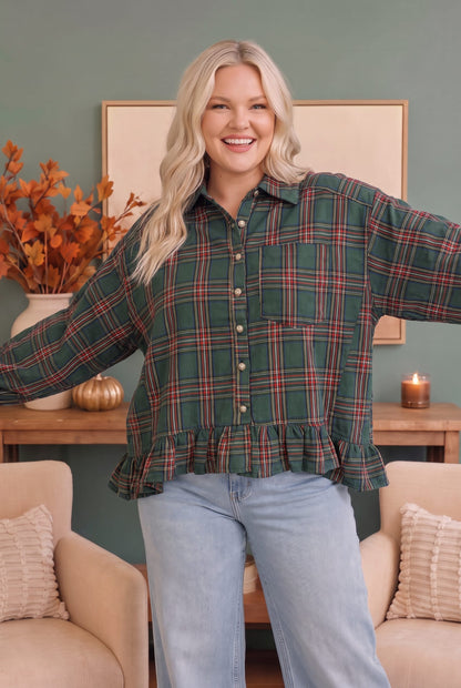 A woman modeling a green plaid button-up shirt with a ruffled hem and light blue jeans, standing in a cozy living room setting with beige armchairs and a wooden console table.