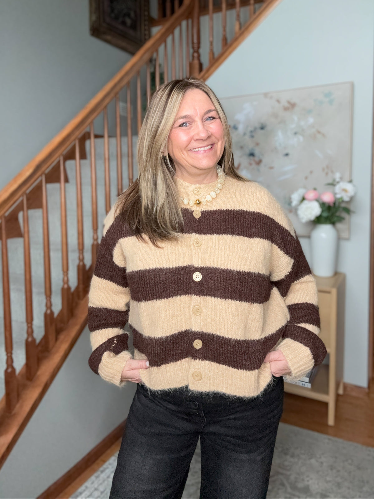A woman smiling in a cozy indoor setting, wearing a brown and beige striped knit cardigan sweater with buttons, paired with dark jeans.