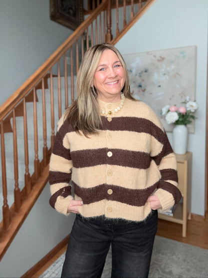 A woman smiling in a cozy indoor setting, wearing a brown and beige striped knit cardigan sweater with buttons, paired with dark jeans.