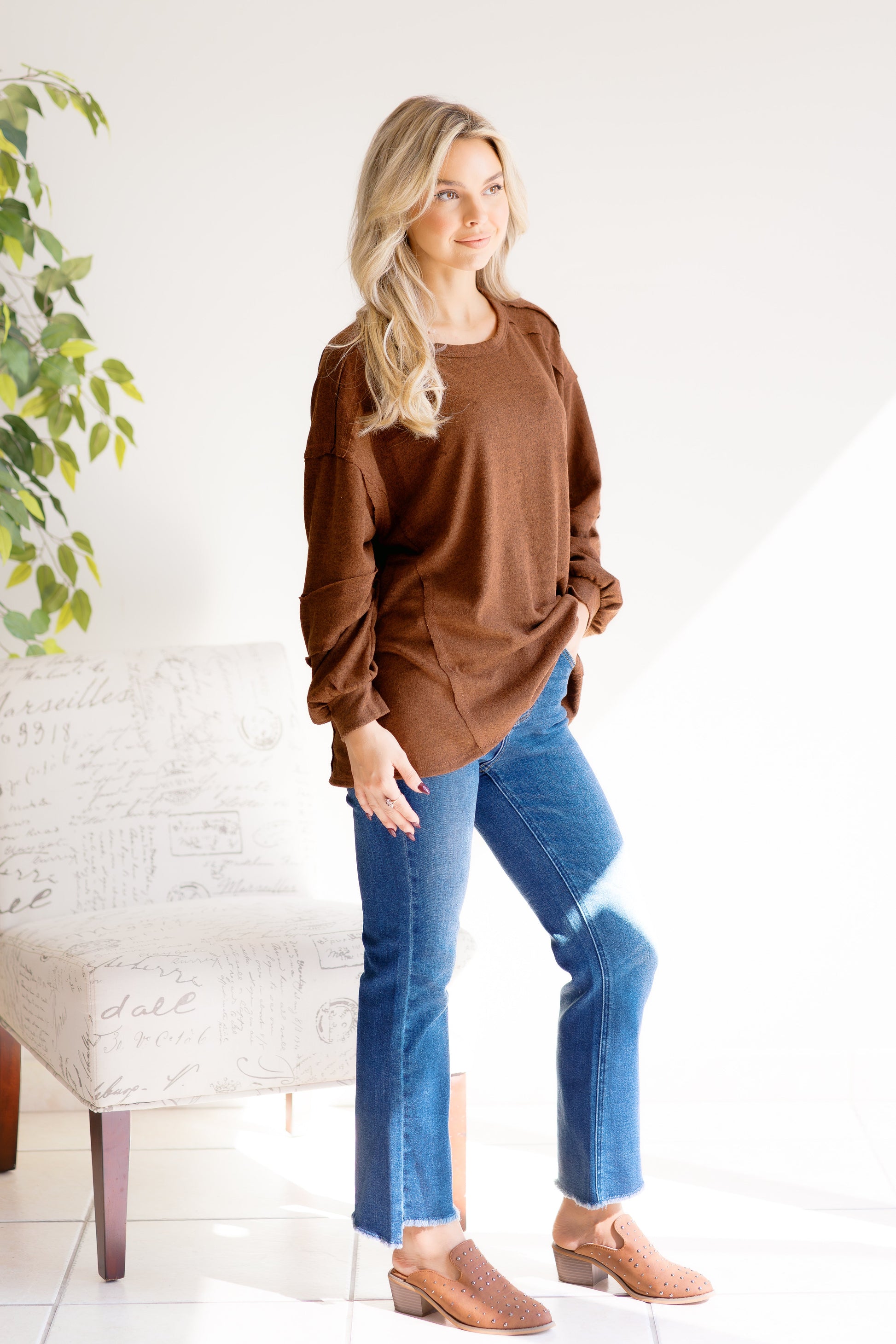 Woman wearing a brown sweater with raw hem and blue jeans standing in a room with a white wall and a plant.