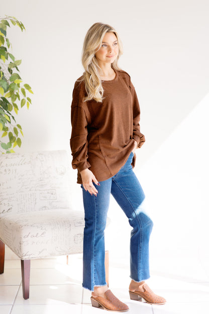 Woman wearing a brown sweater with raw hem and blue jeans standing in a room with a white wall and a plant.