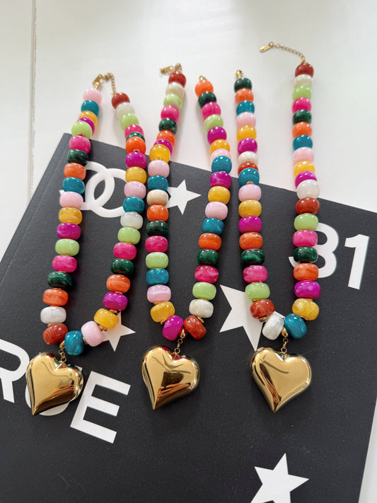 Three colorful beaded necklaces with gold heart pendants, displayed on a black printed card.