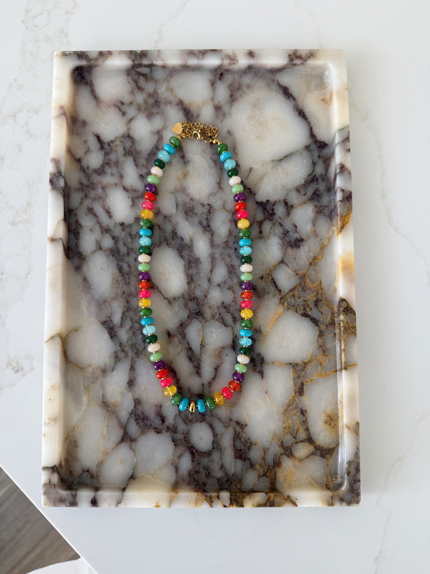 A colorful beaded necklace with a gold clasp, displayed on a rectangular white and gray marble tray.
