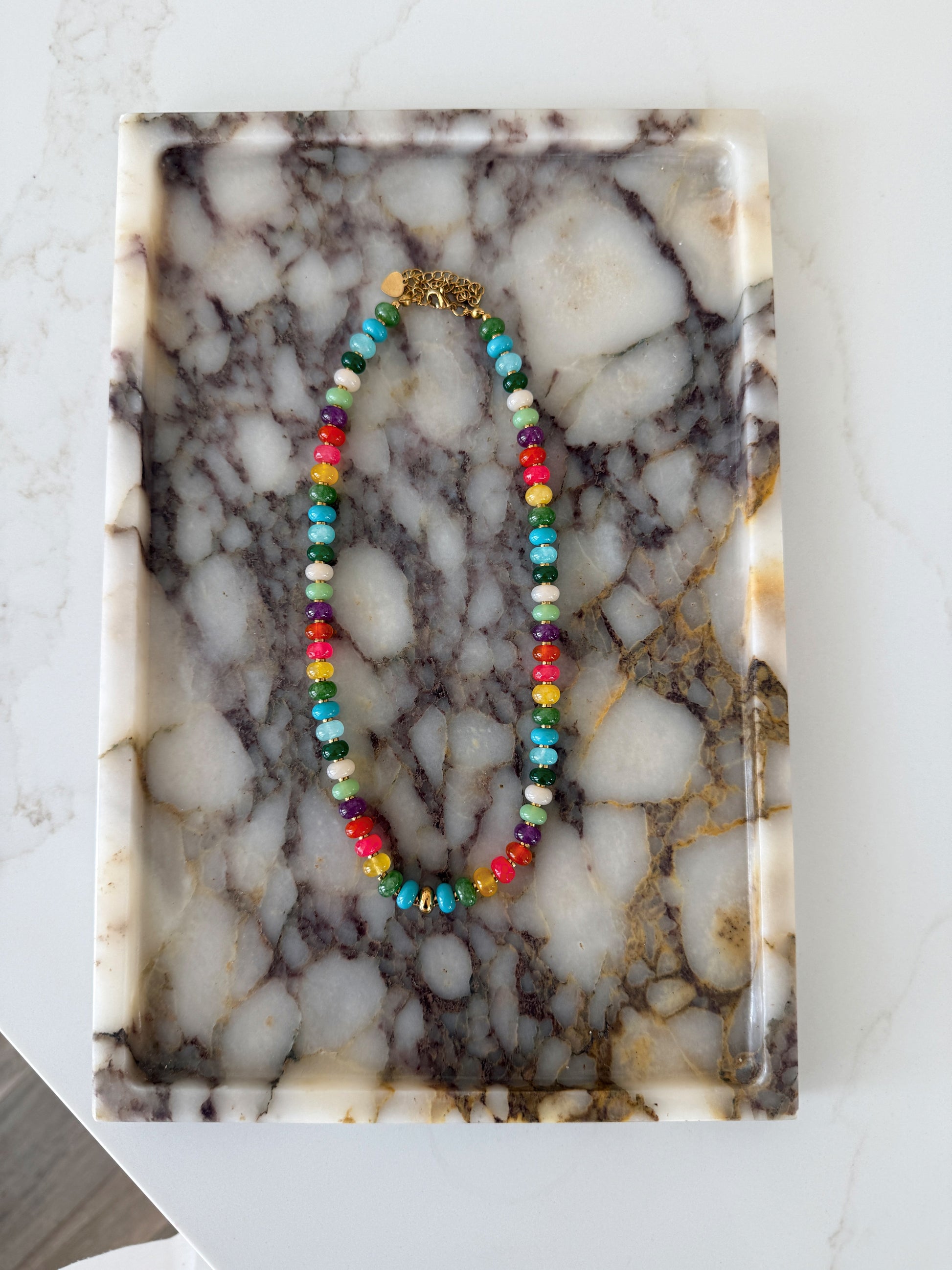 A colorful beaded necklace with a gold clasp, displayed on a rectangular white and gray marble tray.