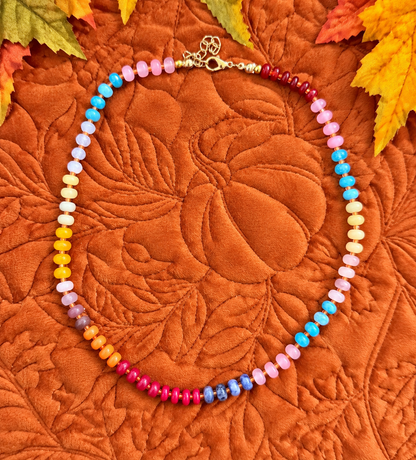 A beaded necklace with a rainbow gradient of colorful round beads and a gold clasp, displayed on a textured orange quilted blanket with autumn leaves in the corners.