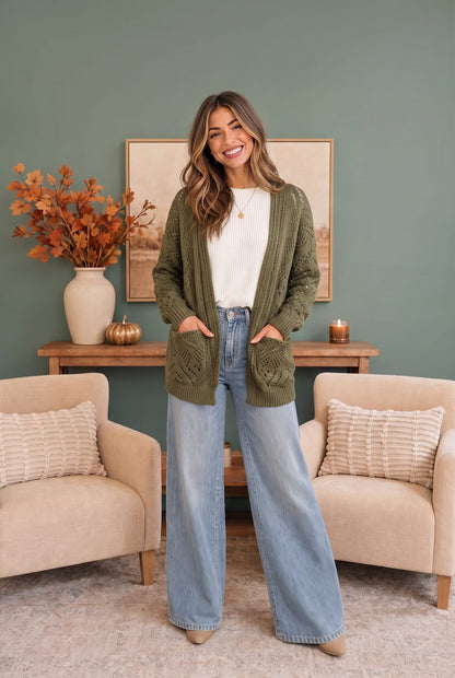 A woman stands in a styled living room wearing an olive green cable-knit cardigan over a white top and light blue wide-leg jeans. The background features a sage green wall, a wooden console table with a vase of autumn leaves, and two beige armchairs.