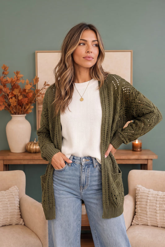 A woman wearing an olive green open-front cable-knit cardigan over a white ribbed top and light blue jeans, standing in a styled room with a sage green wall, beige armchairs, and a wooden console table.