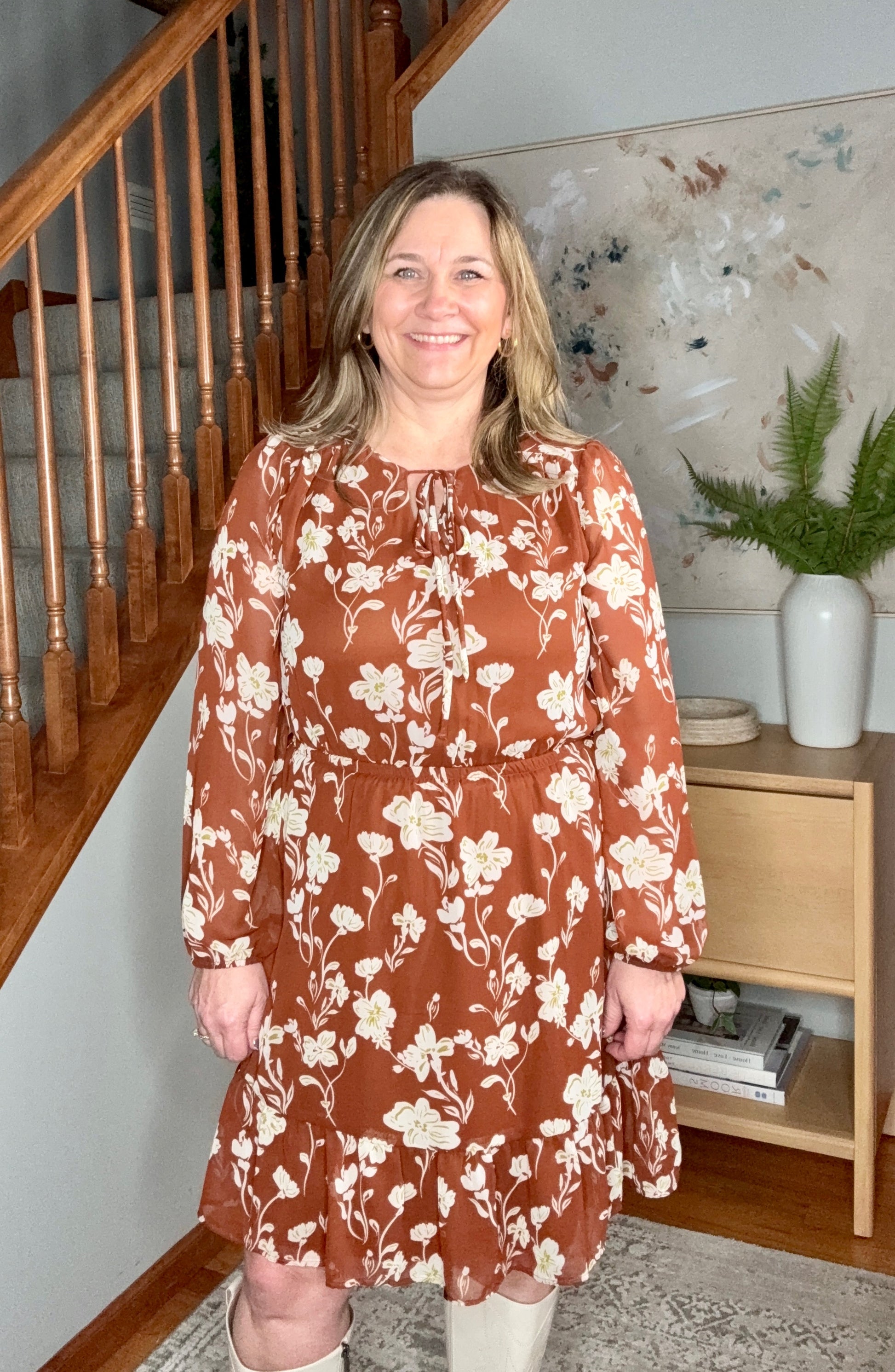 Woman wearing a brown floral dress standing in a home setting with a staircase and decorative items.