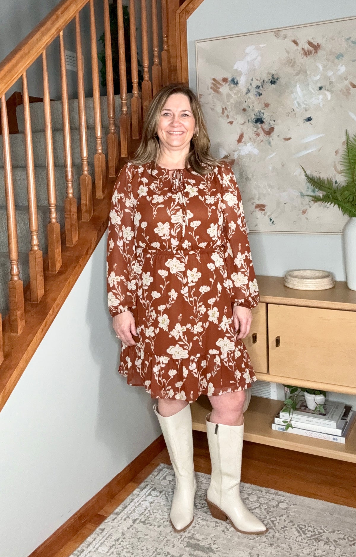 Woman in a brown floral dress and white boots standing in a home interior.