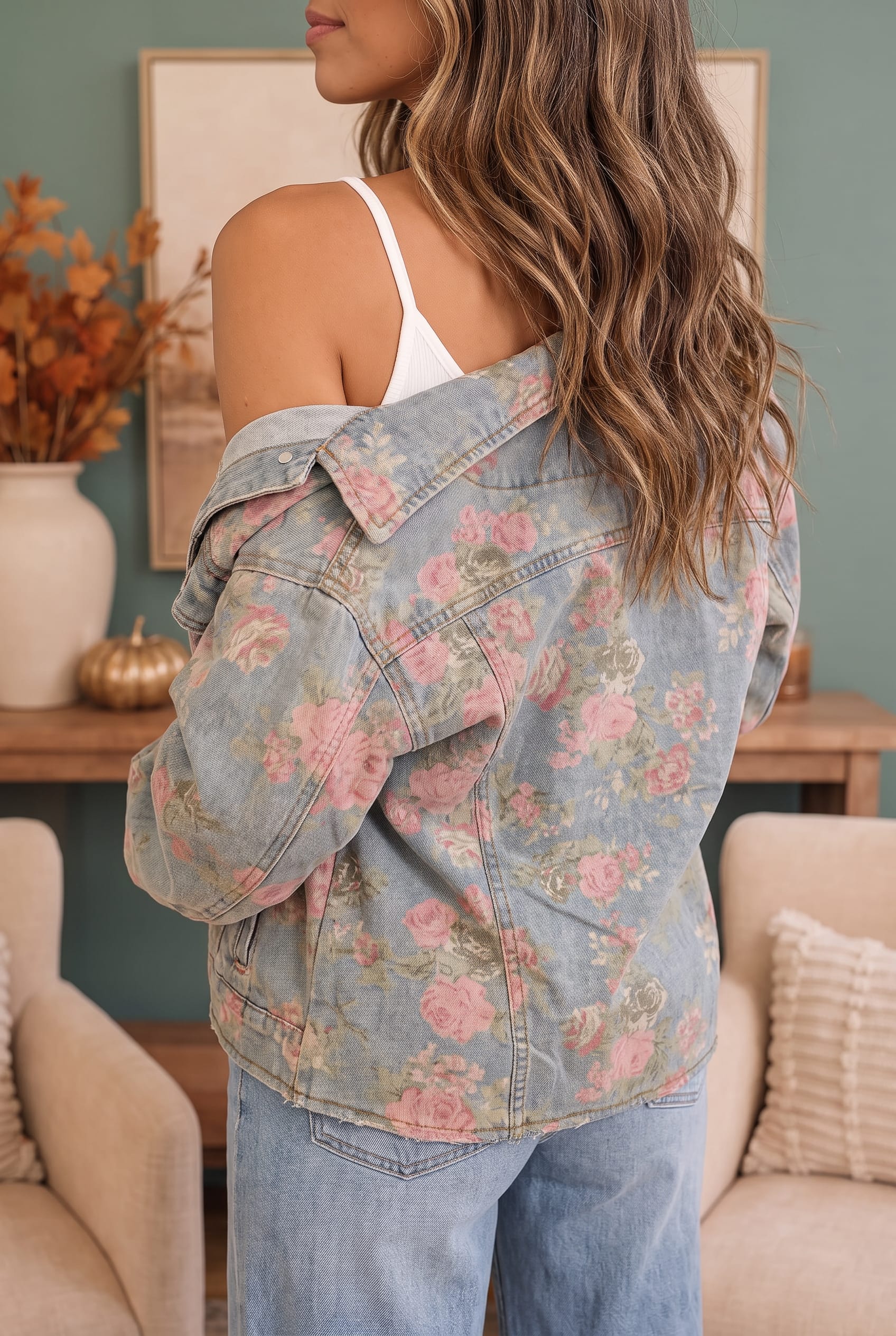 A woman shown from the back wearing a light-wash blue denim jacket with a pink floral print over a white tank top and blue jeans, set in a cozy living room environment.