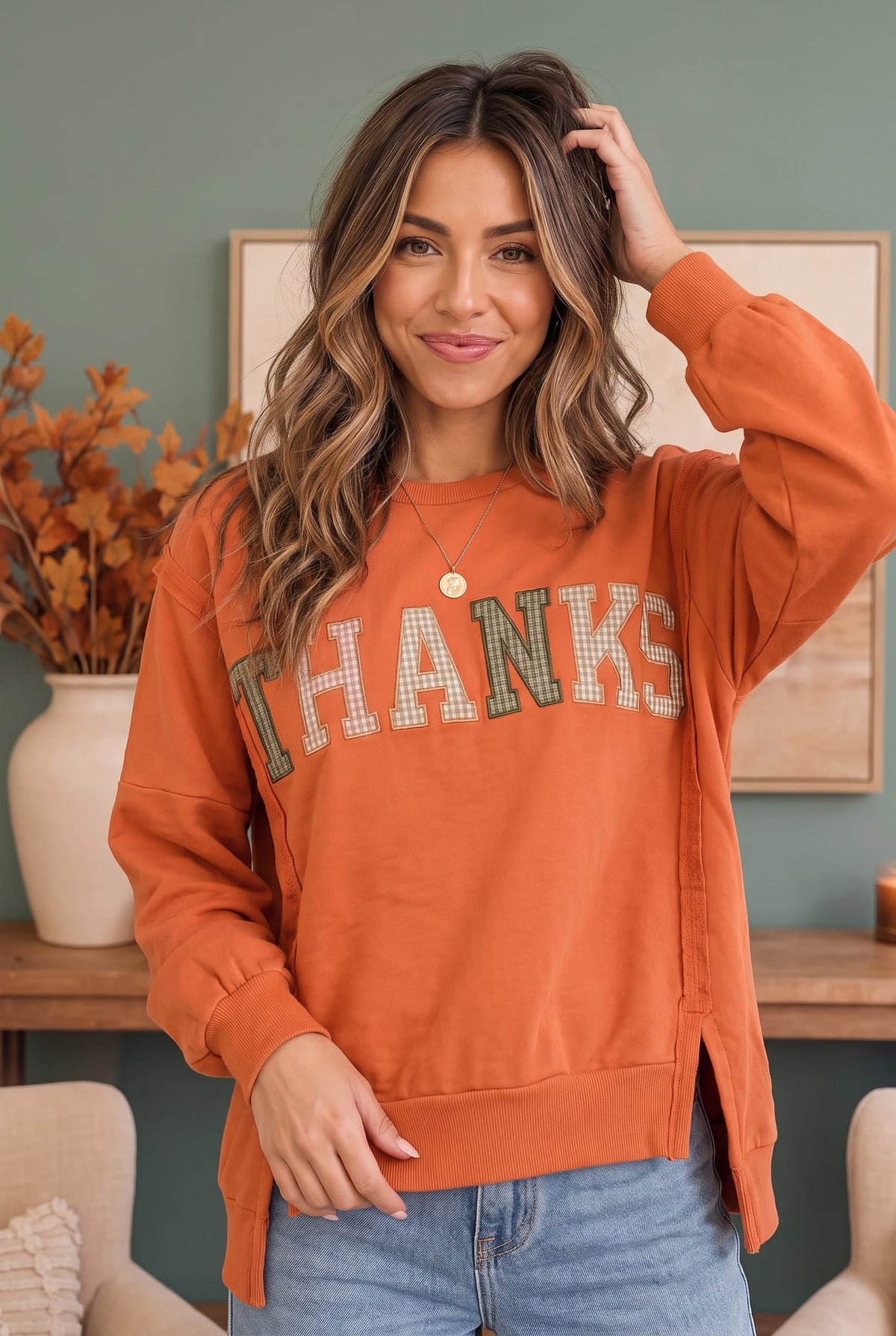 A woman wearing a burnt orange sweatshirt with 'THANKS' printed across the front in a plaid pattern, paired with blue jeans, standing in a cozy indoor setting.