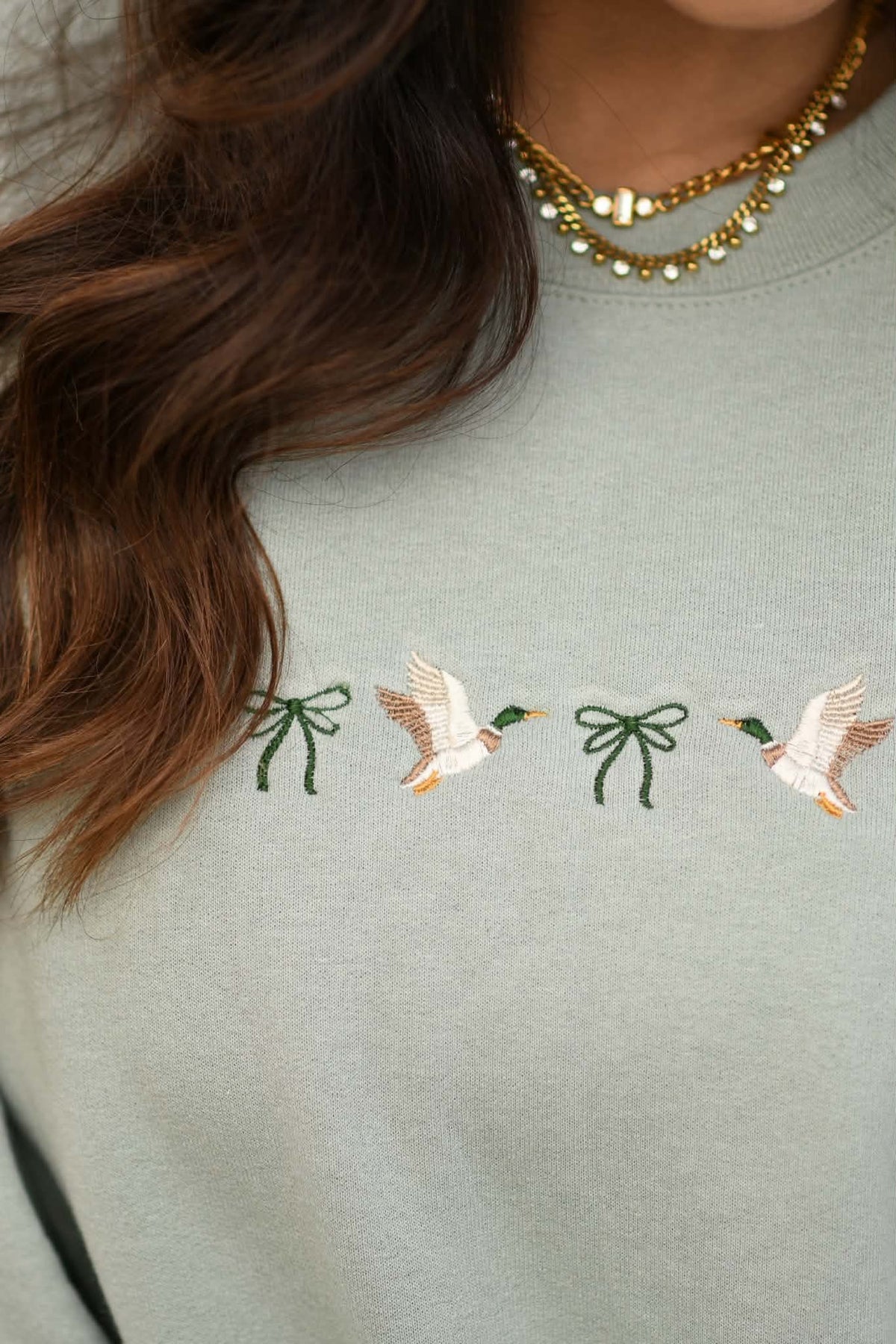 Close-up of a person wearing a sage green crewneck sweatshirt with embroidered mallards and green bows on the chest, accessorized with a gold beaded necklace.