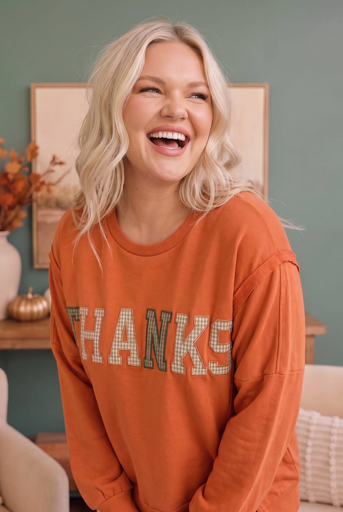 A woman with blonde hair wearing a burnt orange long-sleeve sweatshirt with 'THANKS' written across the front in a gingham pattern, set against a cozy, muted teal living room background.