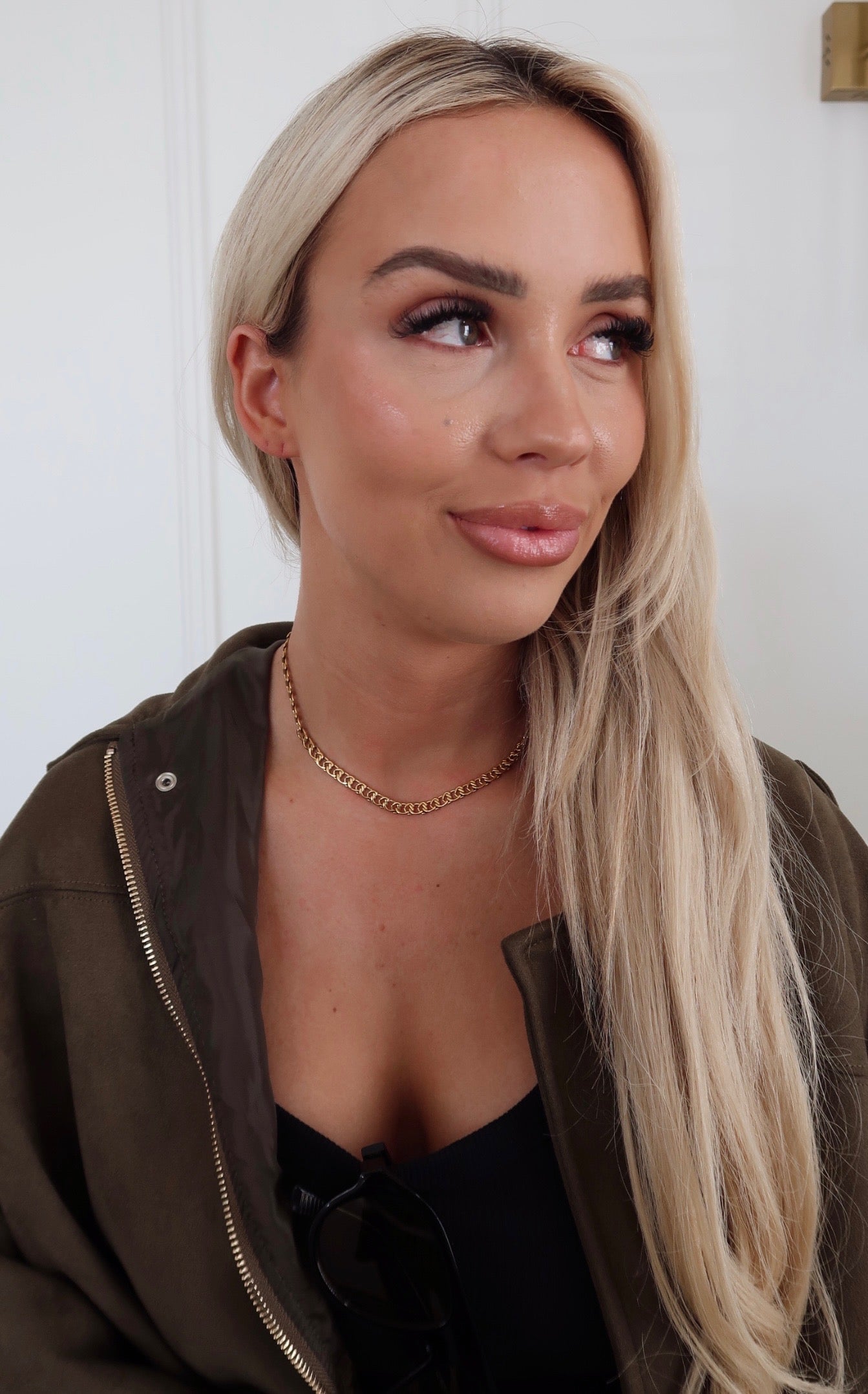 Woman with long blonde hair wearing a brown jacket and black top against a white background
