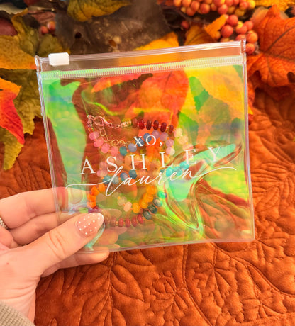 A hand holds a clear zippered pouch with a colorful autumn leaf print and the text 'XO Ashley Lauren' printed on the front. The pouch contains beaded bracelets, and the background features orange autumn leaves and a quilted orange blanket.