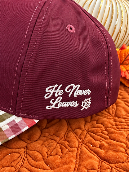 A close-up of a burgundy baseball cap with white embroidered text reading 'He Never Leaves' and a small maple leaf, resting on an orange quilted blanket with a plaid shirt and decorative pumpkins in the background.