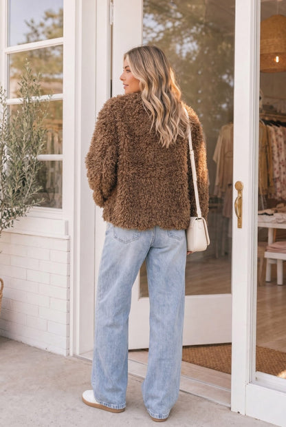 A woman stands in front of a storefront doorway wearing a brown fuzzy textured cropped jacket, light blue wide-leg jeans, and white sneakers, with a small cream shoulder bag.