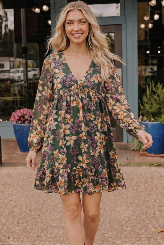 A woman wearing a long-sleeve, V-neck mini dress with a green base and a multicolor floral pattern, standing outdoors in front of a storefront.