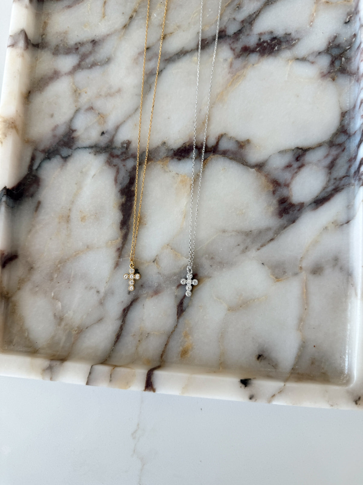 Two delicate cross necklaces, one gold and one silver, displayed on a white marble tray with dark veining.