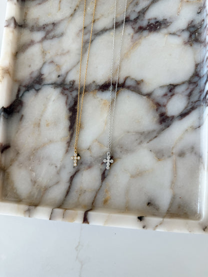 Two delicate cross necklaces, one gold and one silver, displayed on a white marble tray with dark veining.